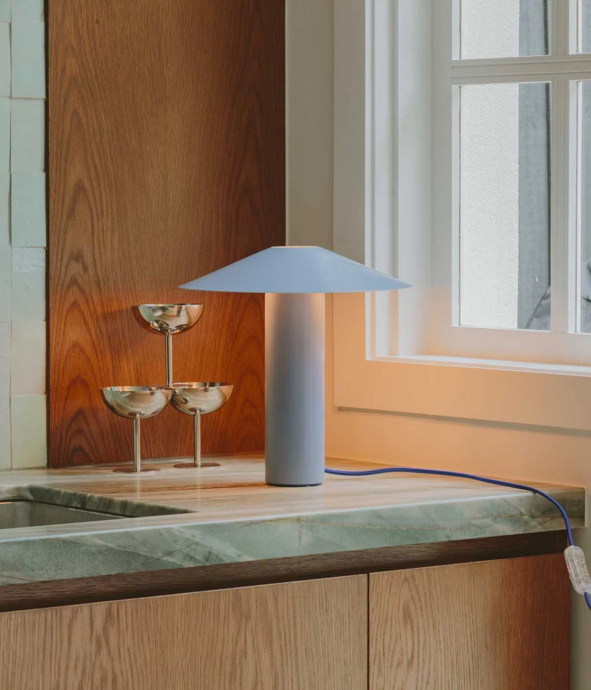 Hoist Lamp in brushed metal on a stone kitchen counter beside silver coupes and warm afternoon window light - Dennis Did It